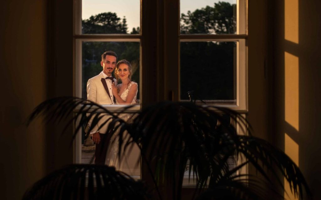 Miri eleganți fotografiați prin fereastră la apus, cu lumină caldă și cadru romantic, fotografie de nuntă artistică realizată de Casa Anke